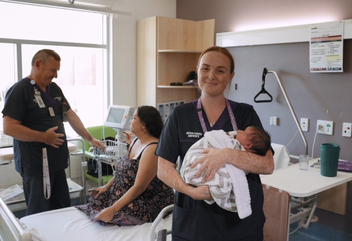 Midwife with baby in arms. Mum and nurse talking in background. 