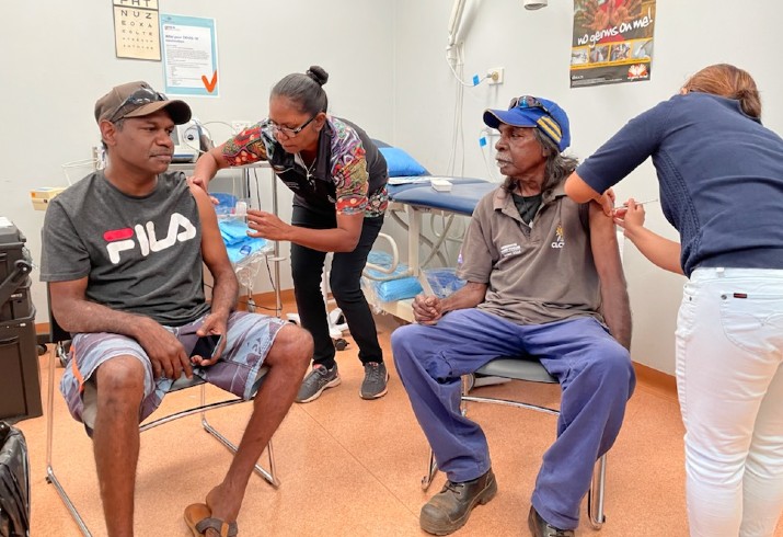 Two men site as two clinicians vaccinate them 