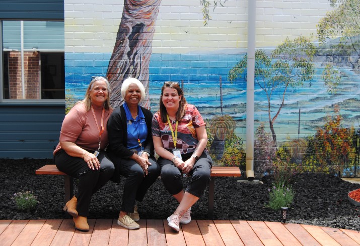 Three staff members sit on bench in garden in front of large mural depicting Australian bush scene