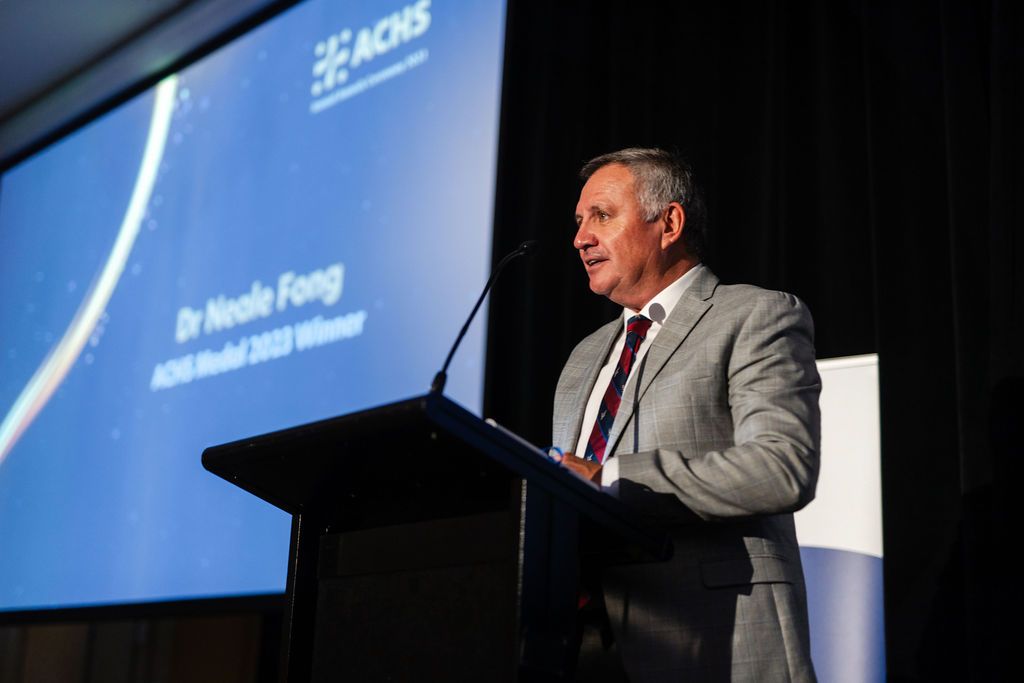 A man stands on stage in front of a screen in a suit speaking to a conference