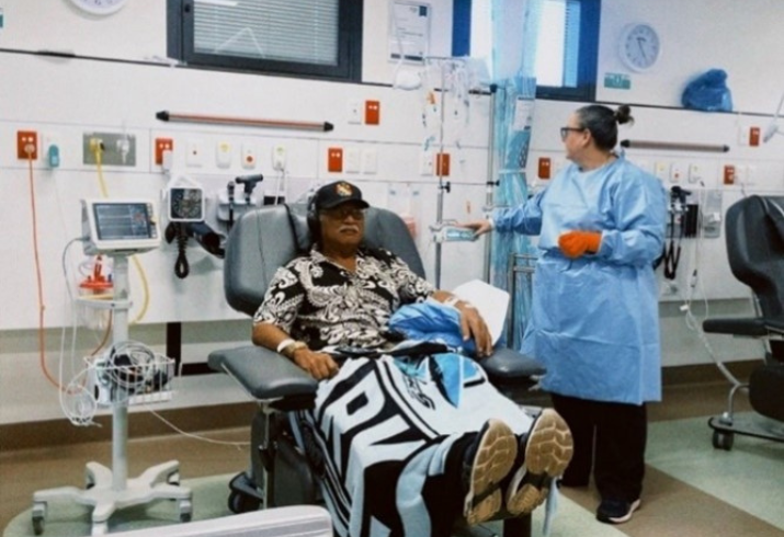 Filipe receiving treatment in hospital chair with Registered Nurse Tracy Hunter standing beside him