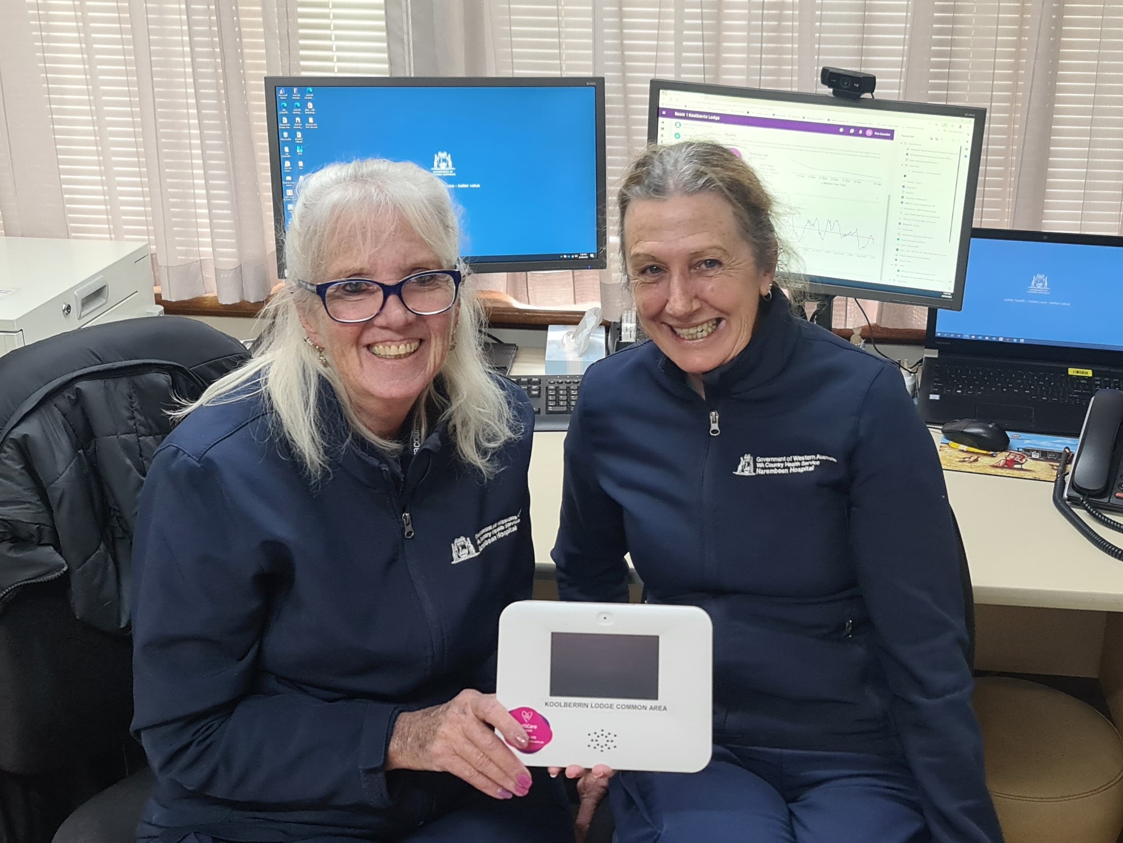 Two healthcare workers hold a digital interface in front of a computer and smile.