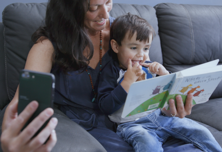 A mum sits with a child on her knee while they read a book, in her other hand she's holding a phone for a virtual child health check