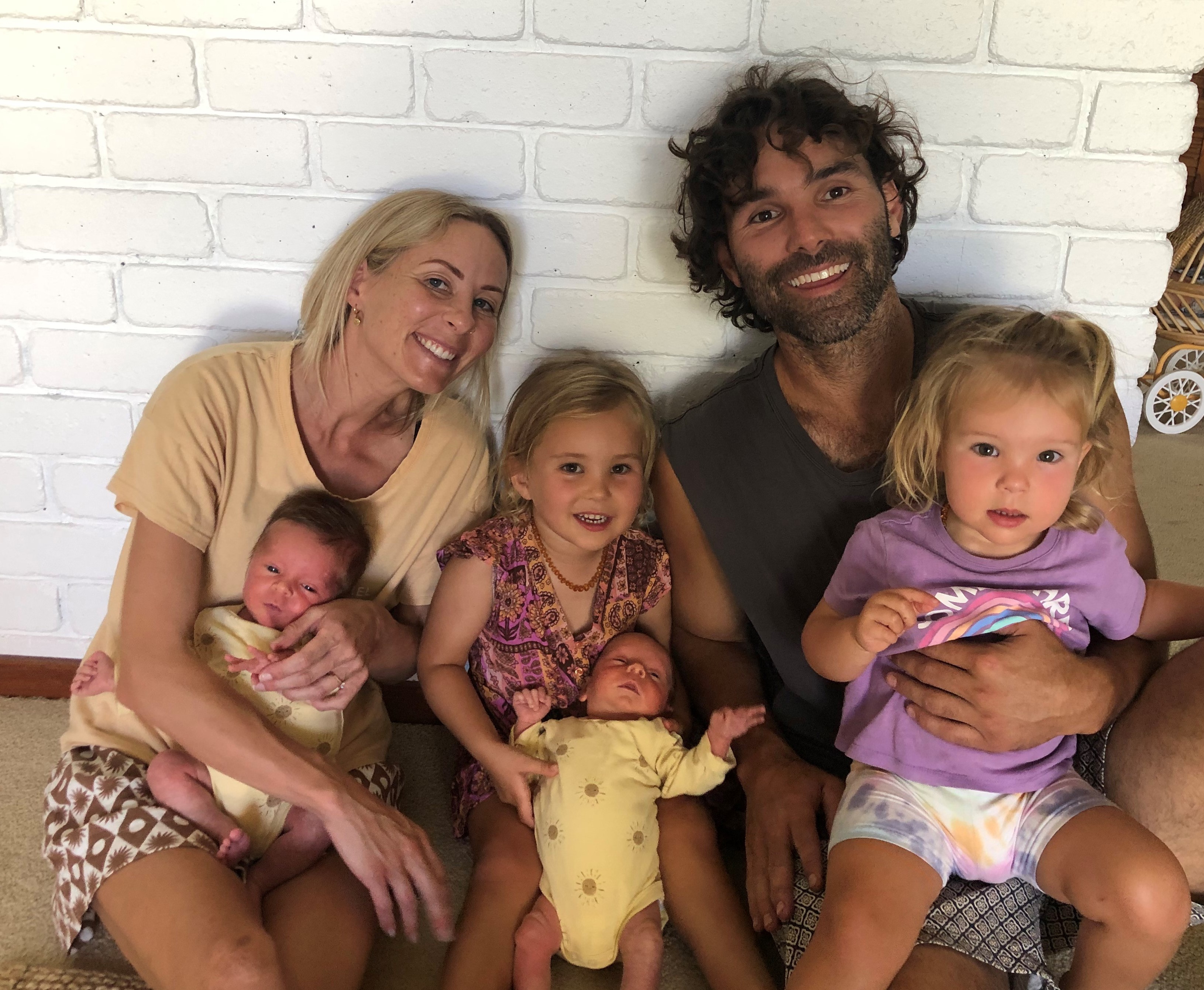 A man and woman sit on the floor agaisnt a wall with four children smiling, two small girls and two infant boys.