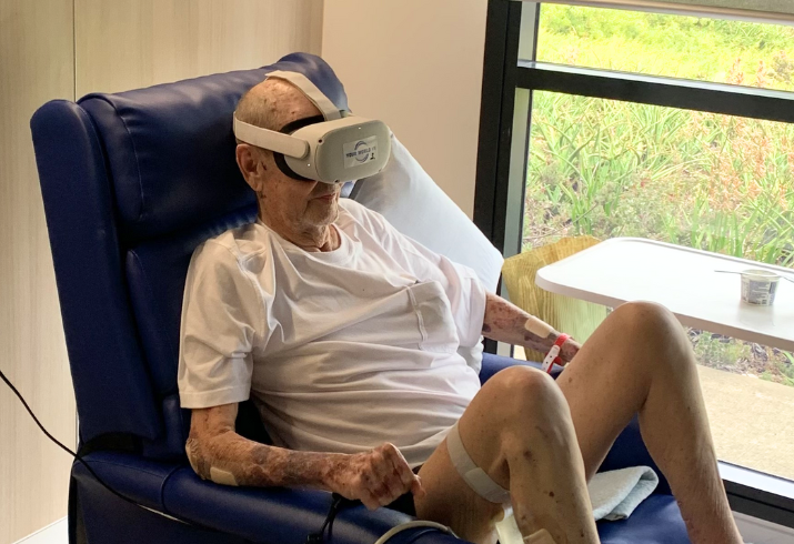 An elderly man uses virtual reality goggles while seated in a chair in a hospital room