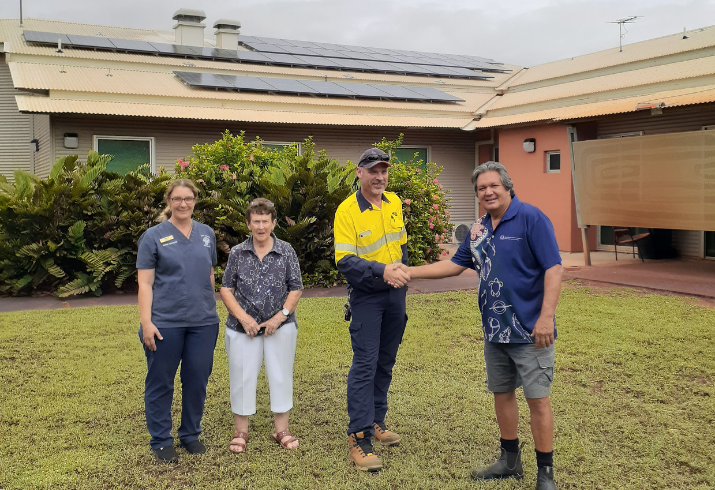 Two women stand by as two men shake hands in front of a hospital building showing new solar panels on the roof