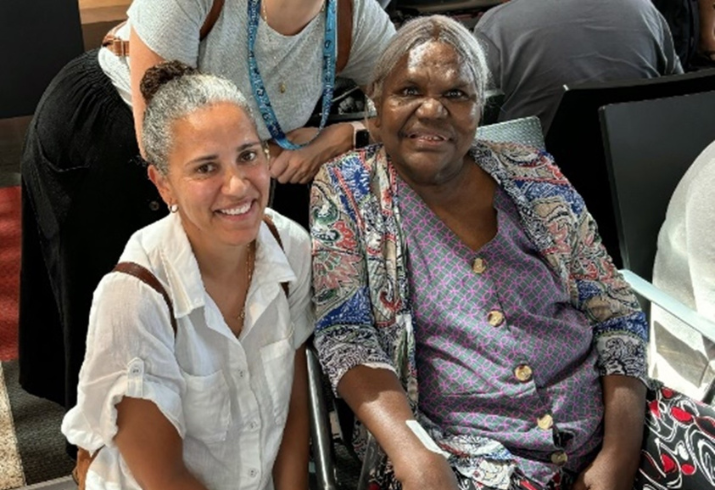 A woman crouches down next to a seated elderly woman, they are both smiling.