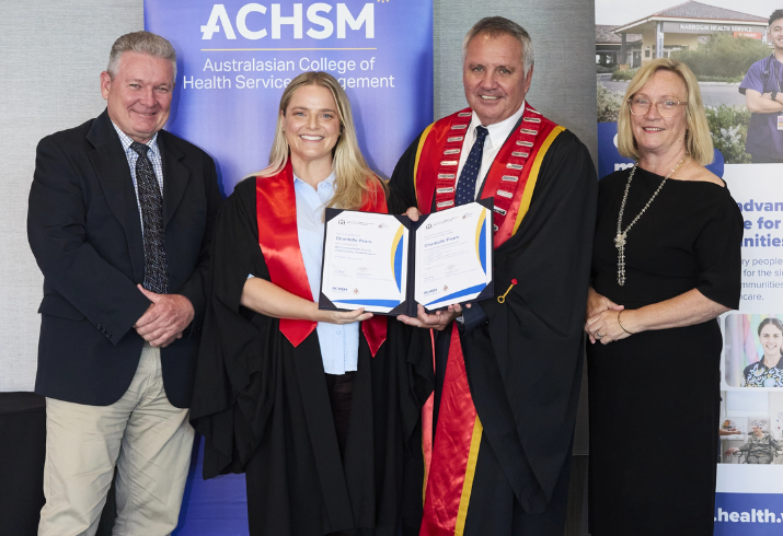 A young woman stands between two men in an academic gown smiling holding an award. Another woman stand to their left also smiling.