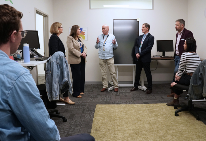 A group of people in an office stand around listening as one man in the centre is explaining to the group