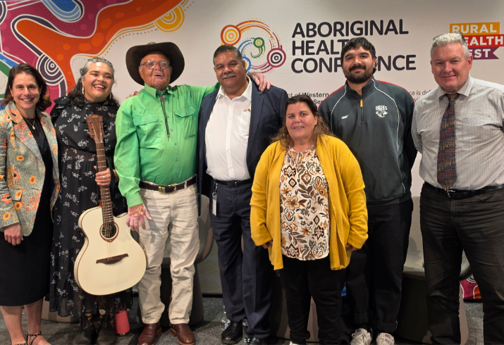 A group of seven people stand on a stage smiling with a backdrop partially obscured that reads Aboriginal Health Conference 2026