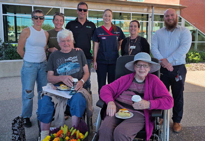 Alan and Glenys sitting in the front row in wheelchairs with a bunch of flowers, cake and coffee, with six hospital staff members standing behind them.