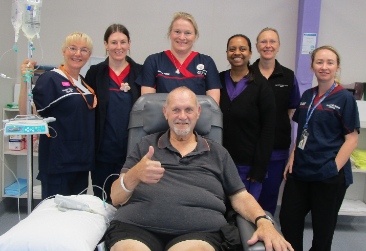 A male patient sitting on a hospital chair with a thumbs up and six nurses behind him all smiling.