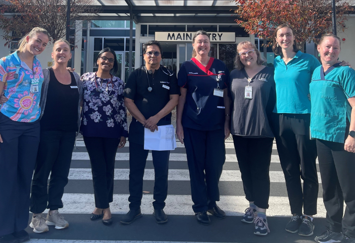 Eight people health uniforms standing in front of hospital entrance.