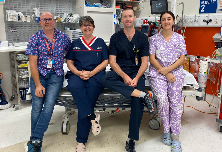 Four health professionals in scrubs sitting on a bed in the hospital.