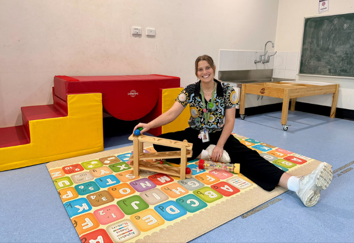 A woman sits on the floor surrounded by brightly coloured play equipment and a rug that features the alphabet