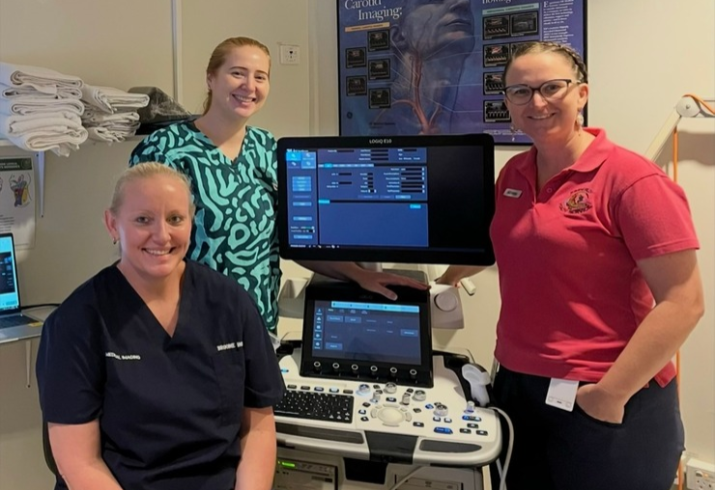 Three women in scrubs stand around a piece of medical equipment smiling
