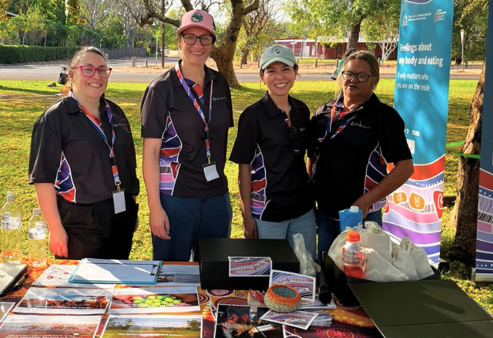 Four women standing behind a table with printed materials behind a colourful banner outside in a park.