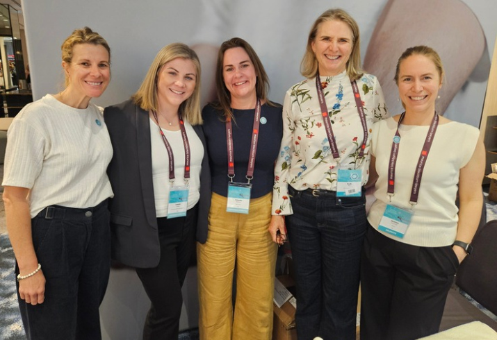 A group of five young to middle-aged women in smart casual attire stand together smiling with conference credentials on lanyards around their necks