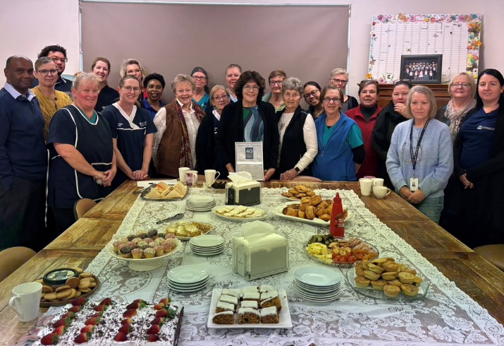 Group of people standing around a table of cakes and food.