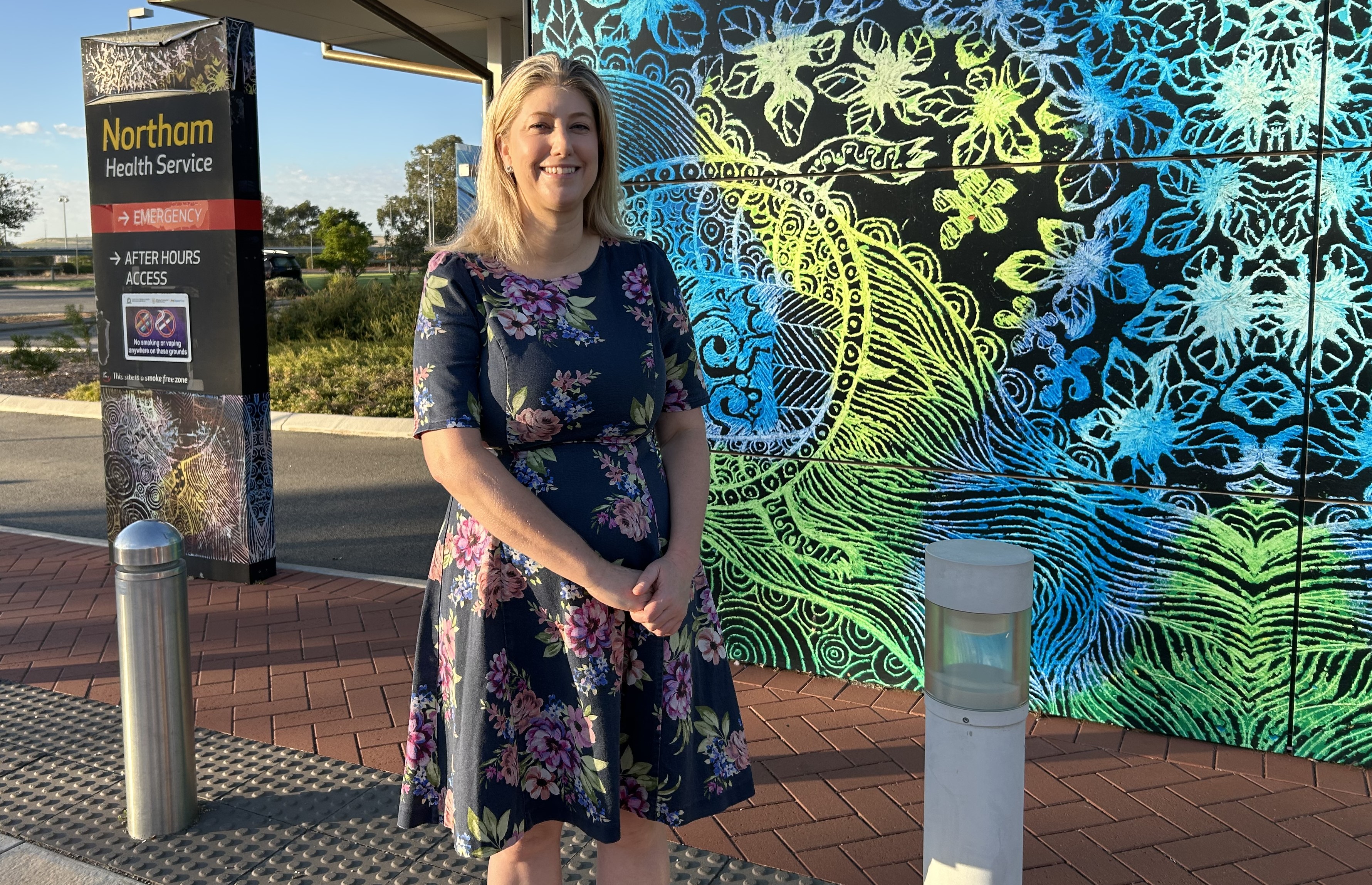 A blond heaired woman stands in front of a brightly coloured screen outdoors, smiling