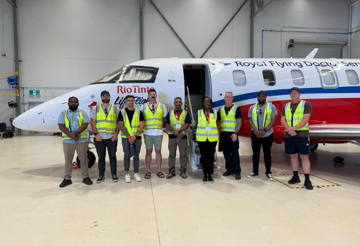 A group of Mental Health Transport Officers stand in front of a Royal Flying Doctor Service plane inside a hangar.