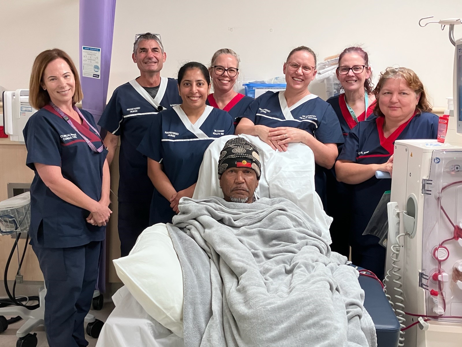A man wrapped in a blanket and seated comfortably receives renal dialysis treatment with a group of seven smiling nurses standing behind his chair