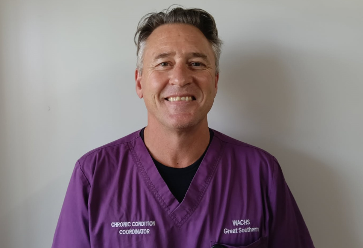Man in purple scrub top with white wall behind.