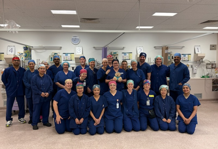 A large group of people in surgical scrubs and caps are gathered around a small teddy bear