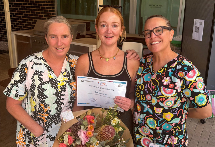 A smiling female nurse with flowers and a certificate stands between two senior nurses in scrubs