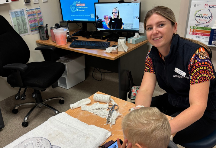A young woman bandages the arm of a small boy facing away from the camera, with another woman visible on screen behind providing clinical support