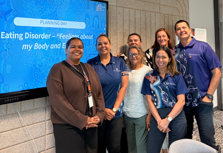 A group of people stand togtehr smiling with a screen behind them that says  Eating Disorder Tool - Feelings about my body and eating