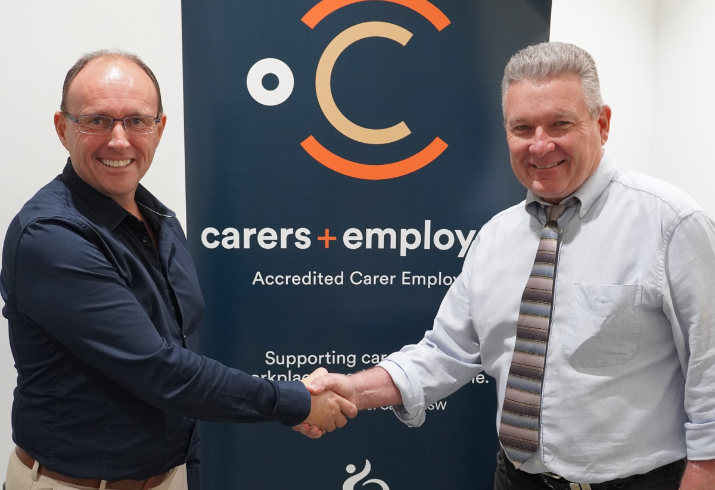 Two men in business casual dress appear on either side of a pull up banner , smile and shake hands.