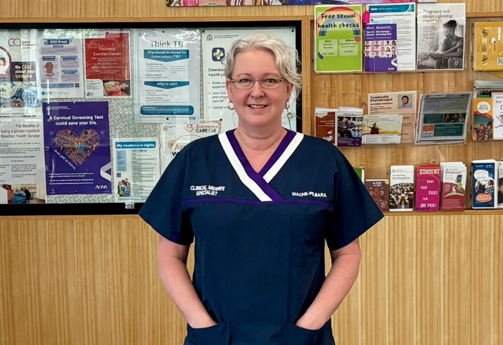 A Clnical Nurse Specialist stands in front of a hospital noticeboard with her hands in her pockets.